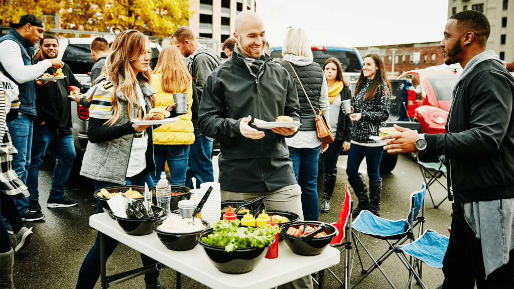 Friends at a tailgate party with food in a parking lot.