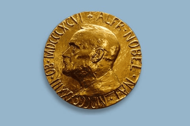 Golden Nobel Prize medal featuring an embossed profile of Alfred Nobel with inscriptions around the edge, set against a plain blue background.