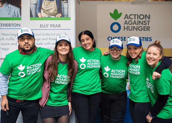 Group of five diverse volunteers wearing green 'Action Against Hunger' t-shirts and caps, smiling and posing together at a charity event booth.